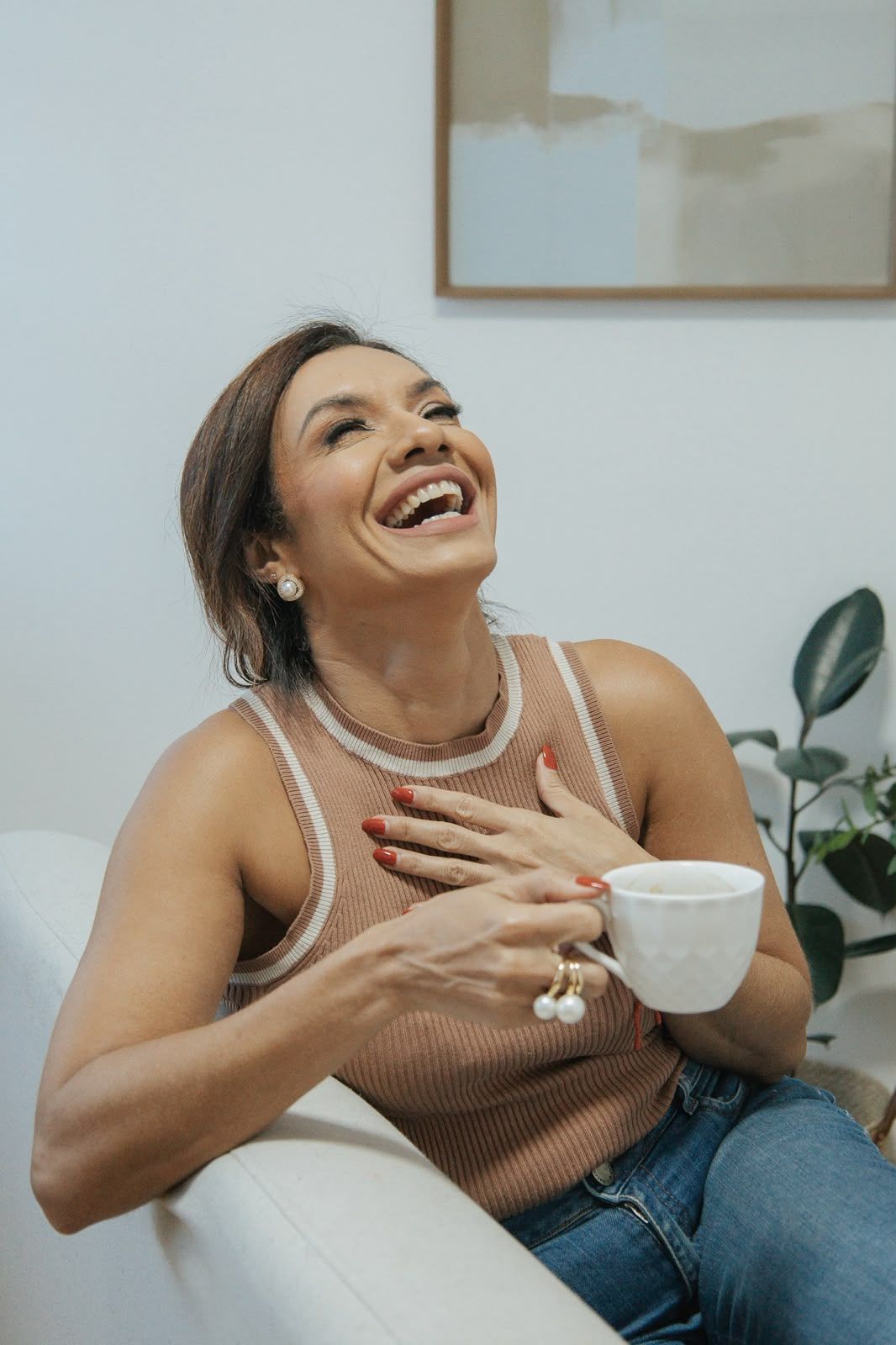 Sandra rindo, segurando uma xícara, sentada em uma poltrona branca